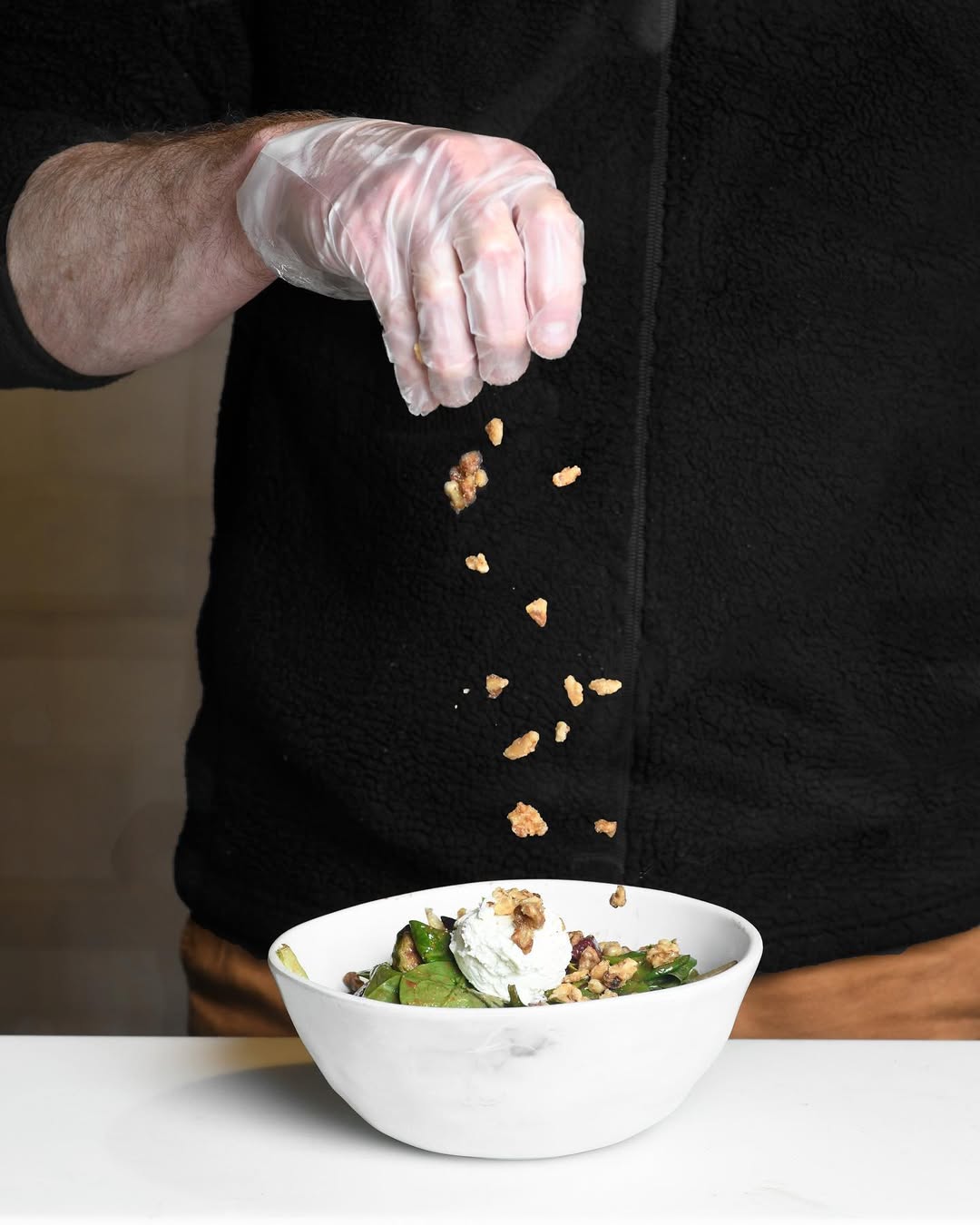 A gloved hand sprinkling crunchy chopped nuts over a fresh salad at Forage Kitchen, adding texture to the greens.