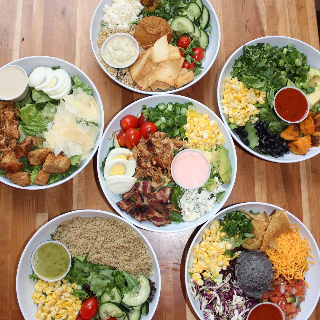 Overhead view of six colorful Forage Kitchen bowls arranged in a circle on a wooden table, filled with vibrant grains, greens, and toppings.