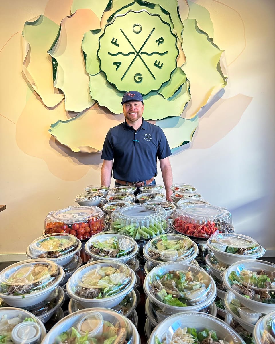 A Forage Kitchen employee standing behind a large catering order of neatly stacked, pre-packaged salad and grain bowls ready for delivery.
