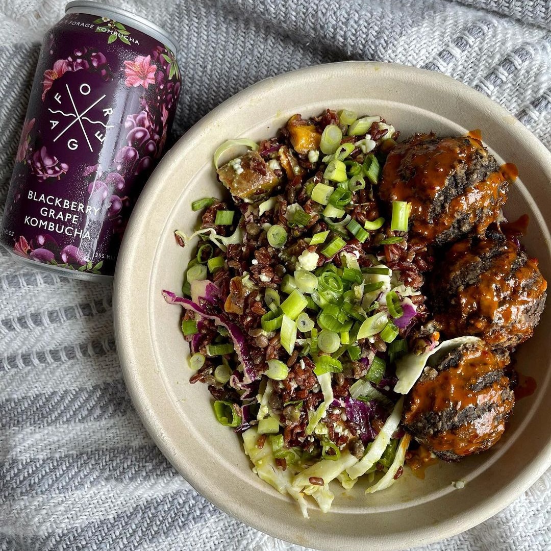 A Forage Kitchen vegan Power Bowl with lentil meatballs and green onion, served beside a glass of kombucha on a cloth-covered table.