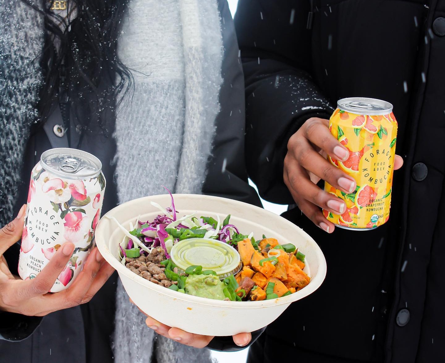 A couple holding a Forage Kitchen grain bowl and kombucha cans in a snowy place.