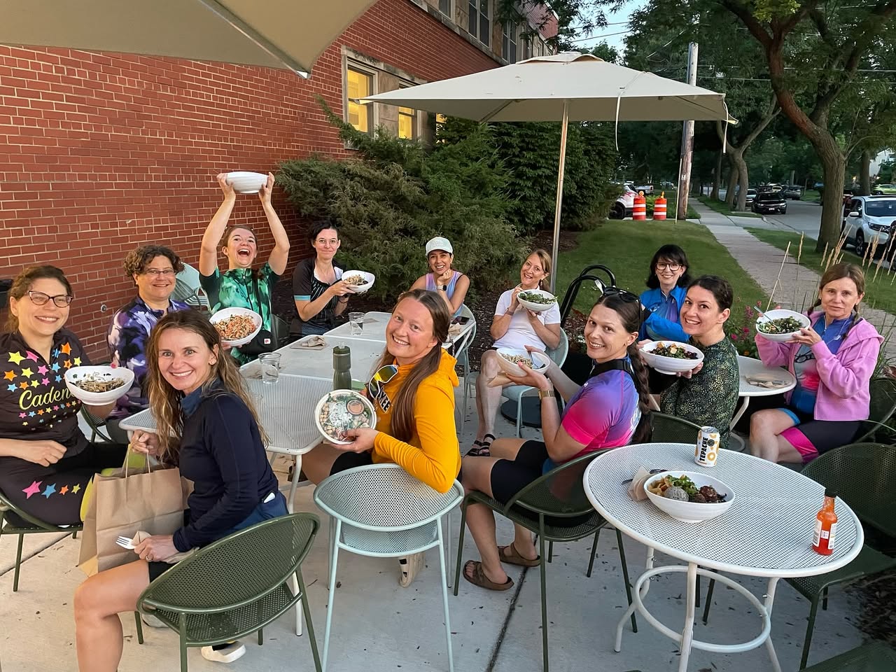 A large group of women in cycling outfits smiling and holding Forage Kitchen grain bowls at outdoor patio tables in Whitefish Bay.