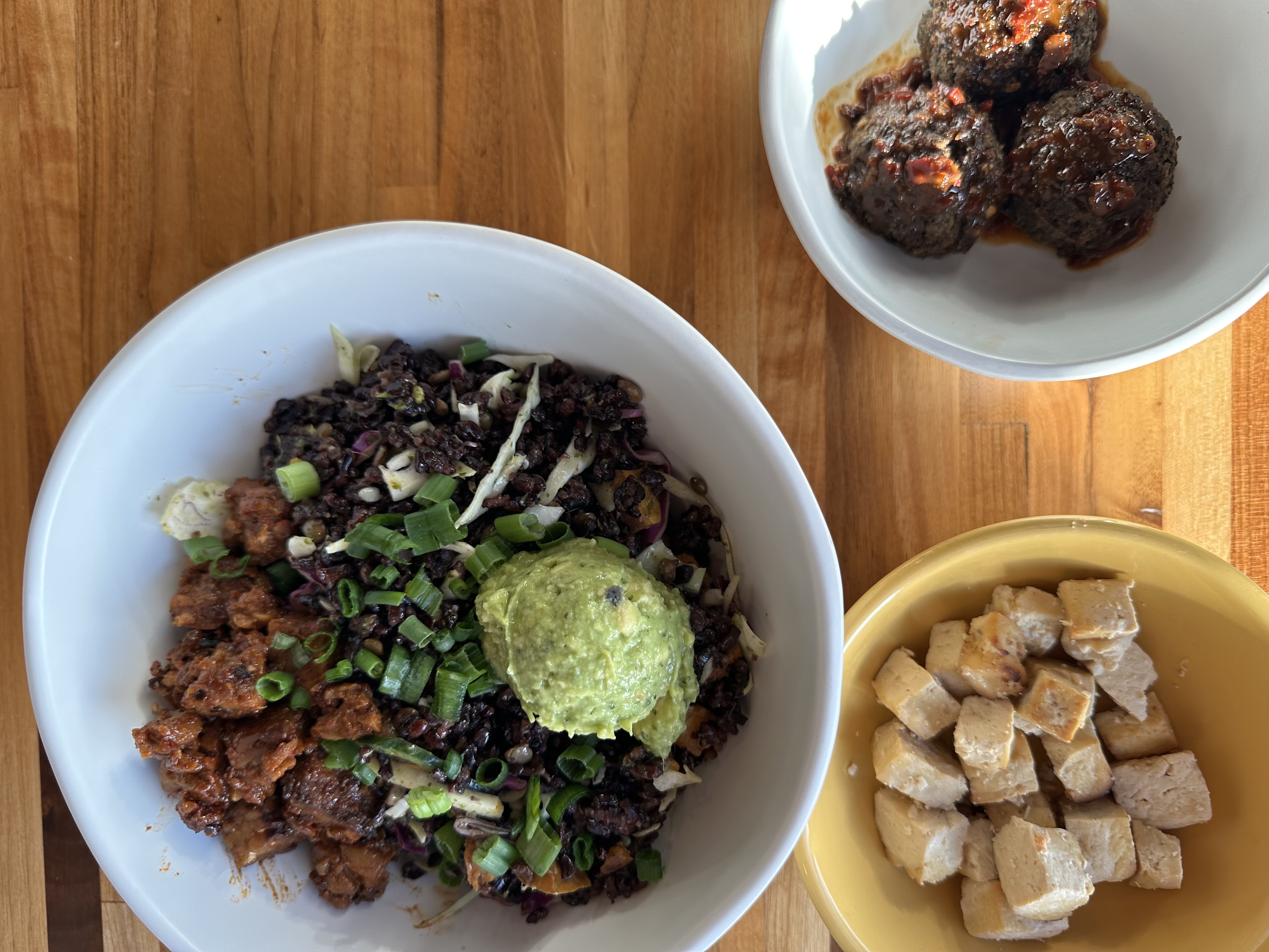 Overhead view of a vegan grain bowl with black rice, guacamole, roasted tofu, and lentil meatballs arranged in sections.