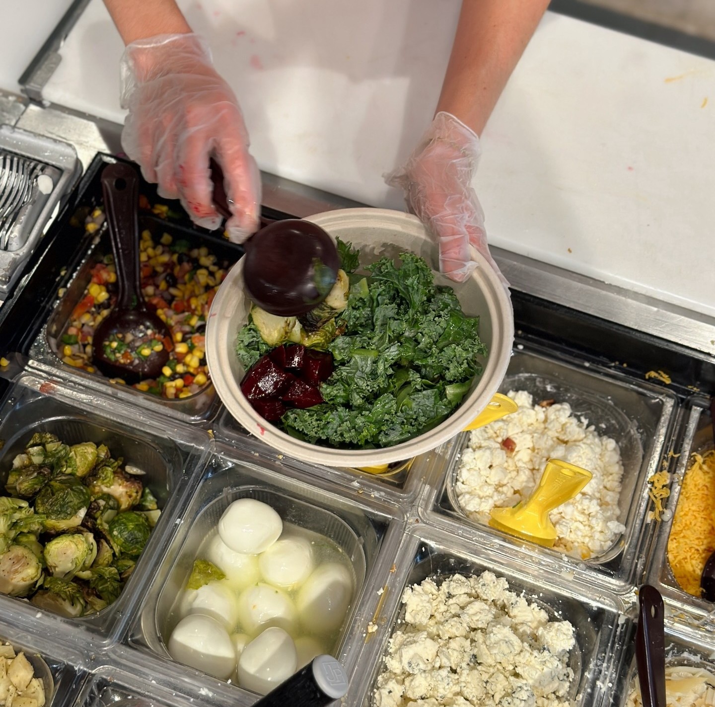 A gloved hand builds a salad with fresh kale and beets at a food prep station.