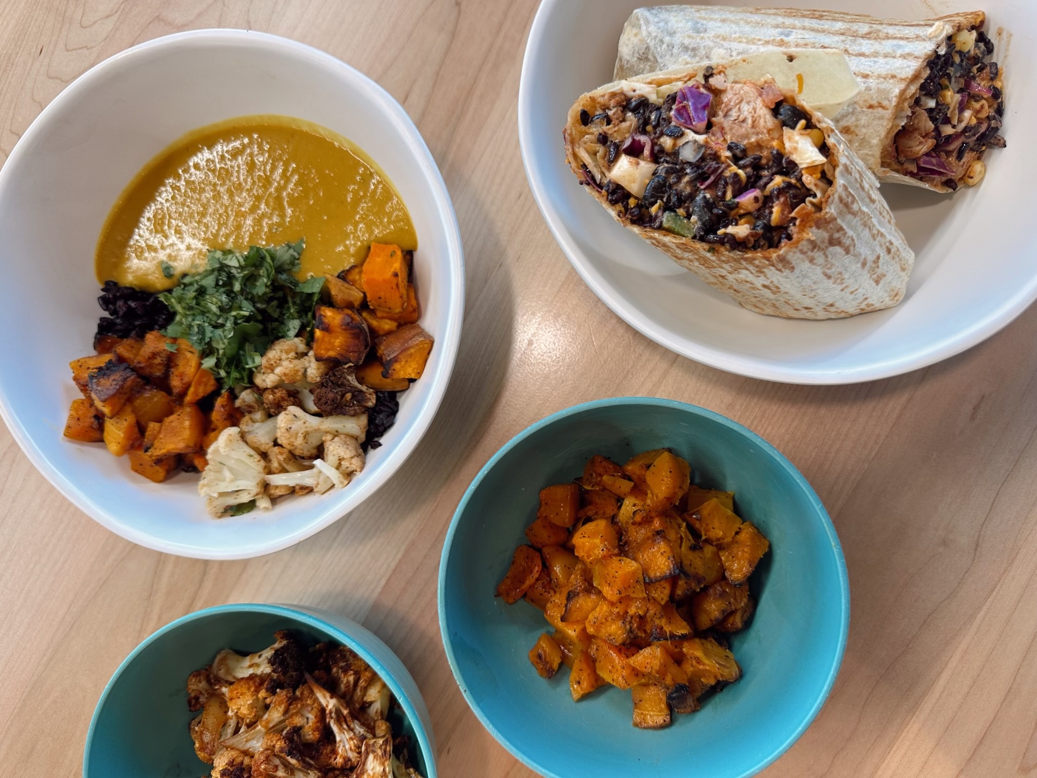 Forage Kitchen Thai bowl, wraps, roasted cauliflower, and sweet potatoes served in colorful bowls on a wooden table.