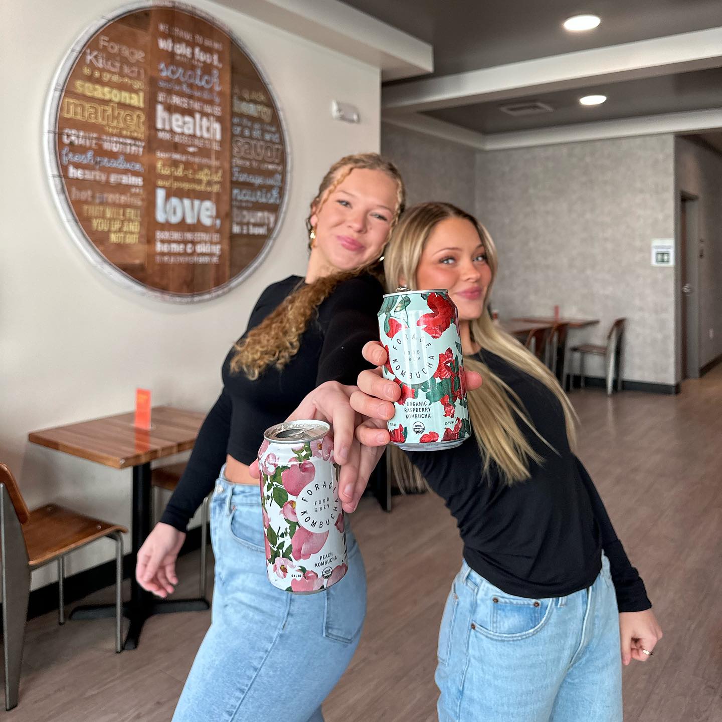 Two women twinning in black tops and blue jeans holding cans of Forage kombucha, smiling and posing.