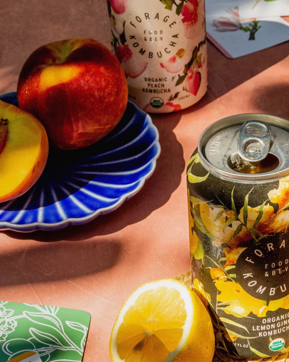 Forage Kitchen Organic Peach and Lemon-Ginger kombucha cans with freshly cut fruits on a table.