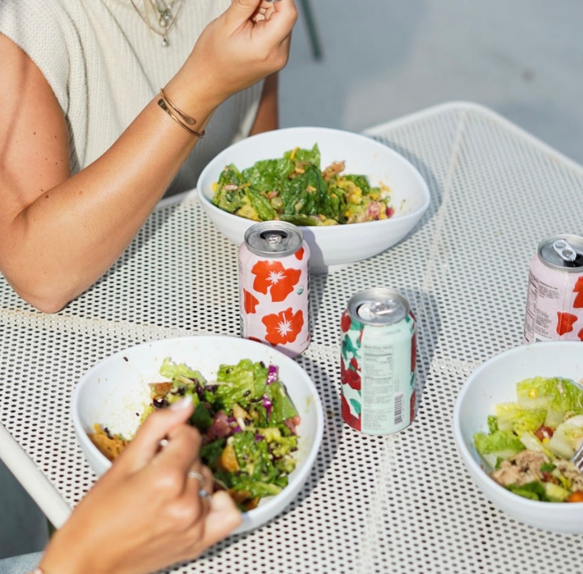  Three people eating fresh Forage Kitchen salad bowls, with cans of kombucha and tepache on an outdoor table.