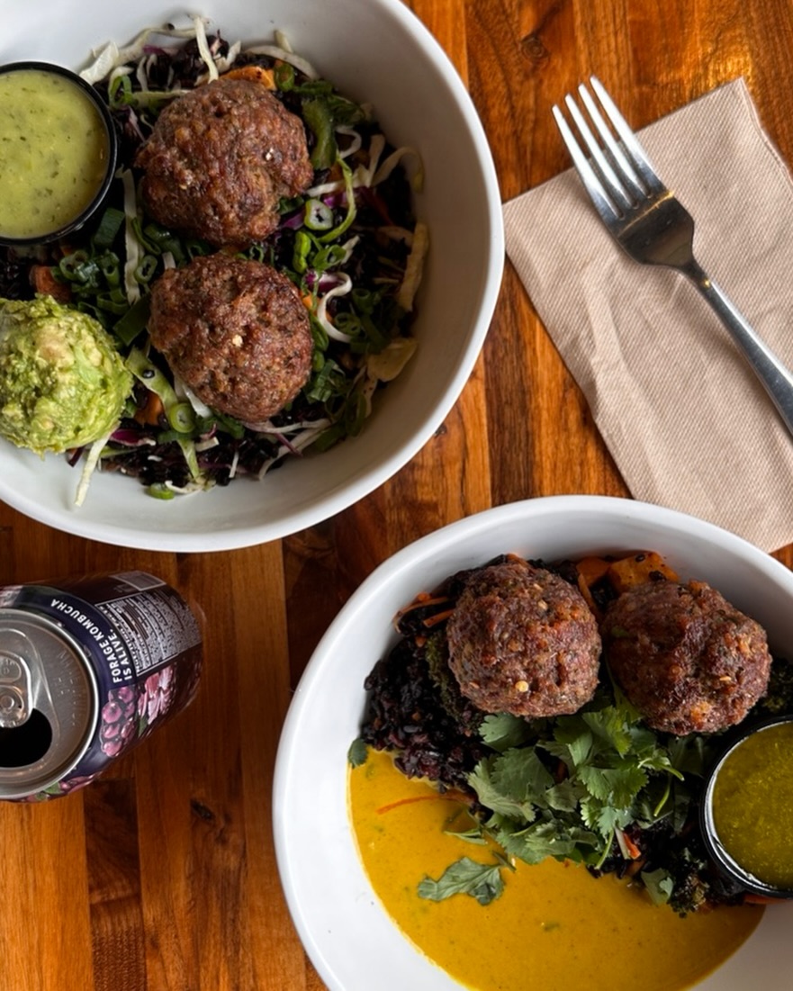 Forage Kitchen Power Bowl and Thai Bowl beside a can of Forage kombucha, a napkin and fork on a wooden table.