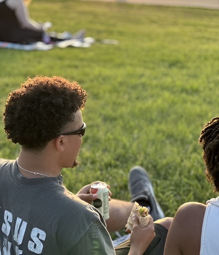 Two people enjoy a wrap and a drink from Forage Kitchen while relaxing on a sunny grassy field.