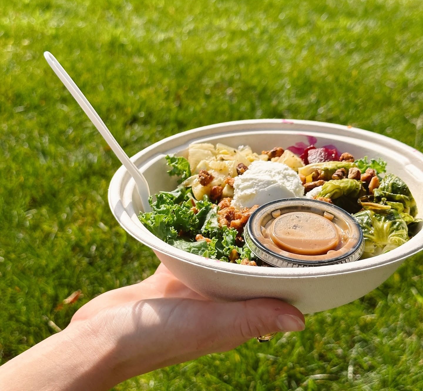 A person holds a fresh Forage Kitchen grain bowl filled with kale and vegetables over a lush green grass field.