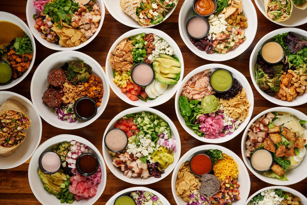 Overhead view of multiple colorful, diverse grain and salad bowls on a wooden table at Forage Kitchen.