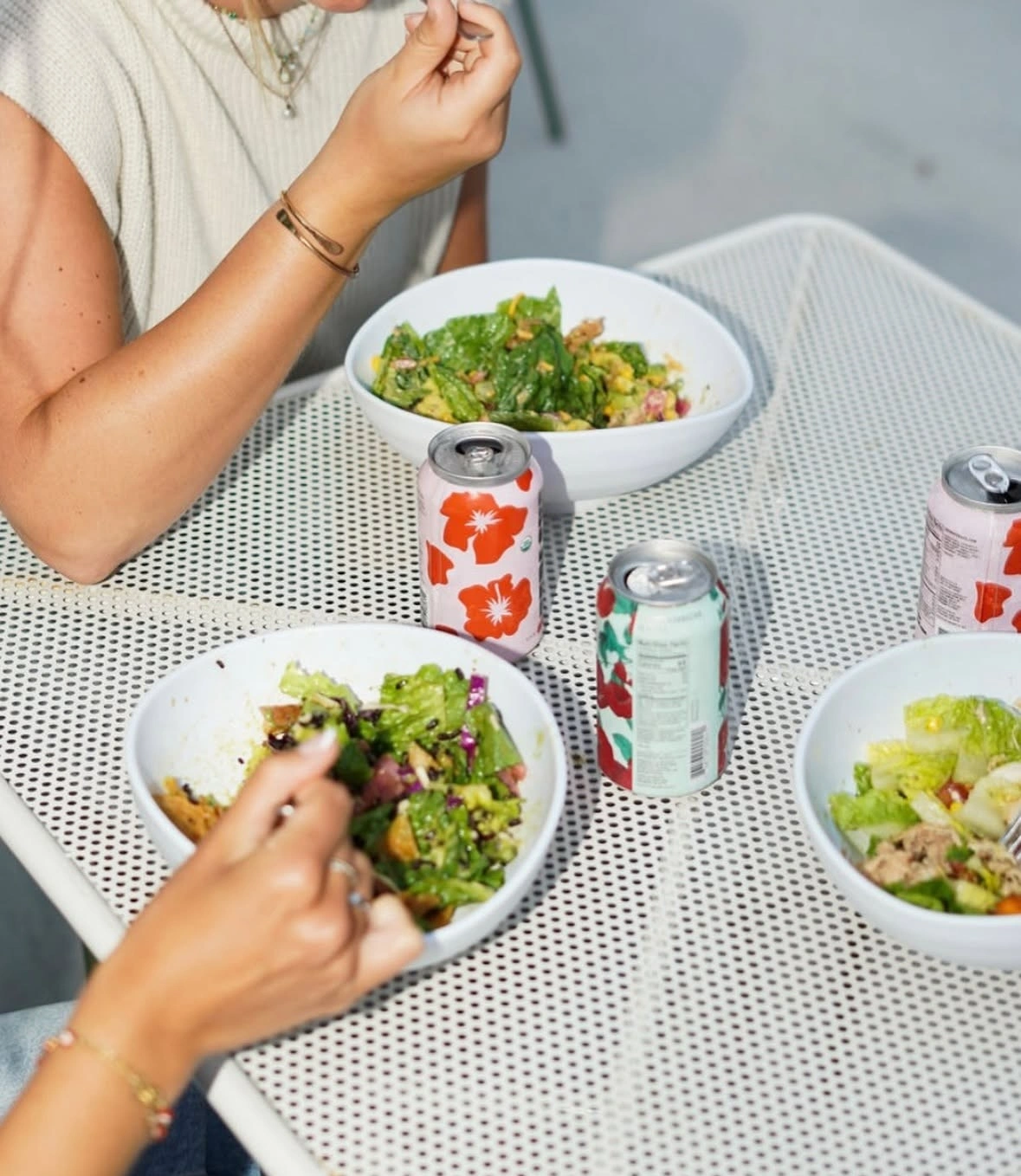 People eating fresh salad bowls with cans of Forage tepache and kombucha at a white Forage Kitchen patio table.