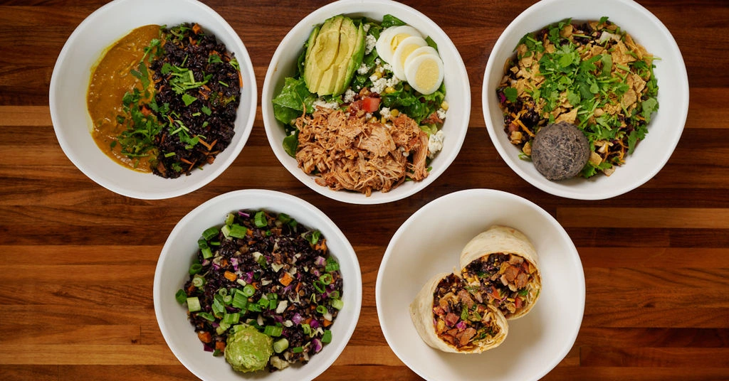 Forage Kitchen grain bowls, salads, and wraps in five white bowls on a wooden table.