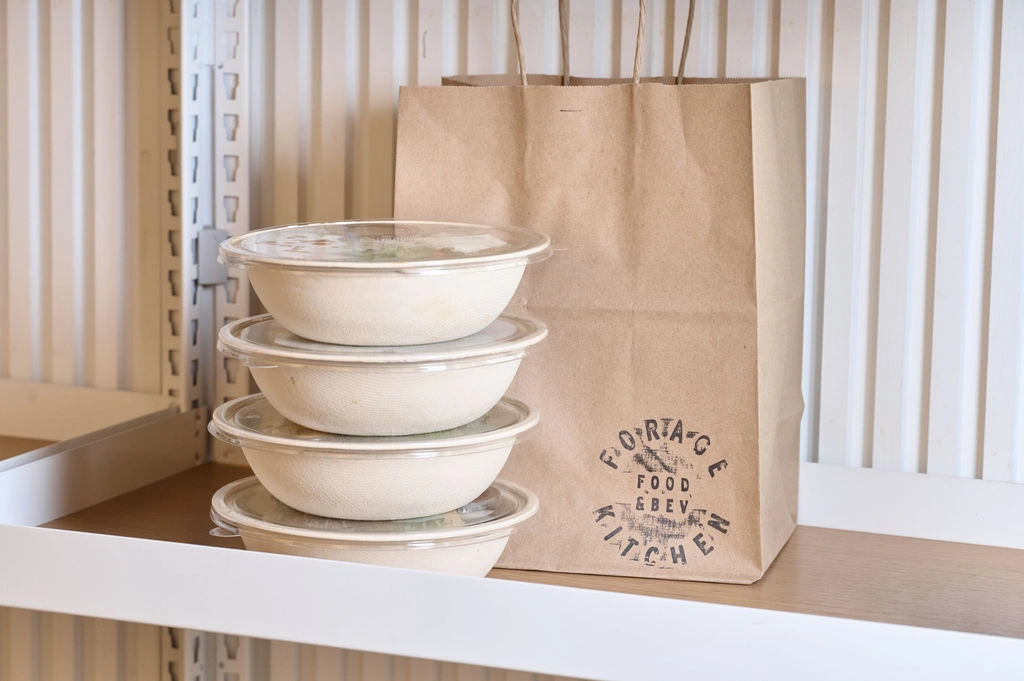 Stacked takeout bowls next to a stamped takeout paper bag on a shelf at Forage Kitchen.