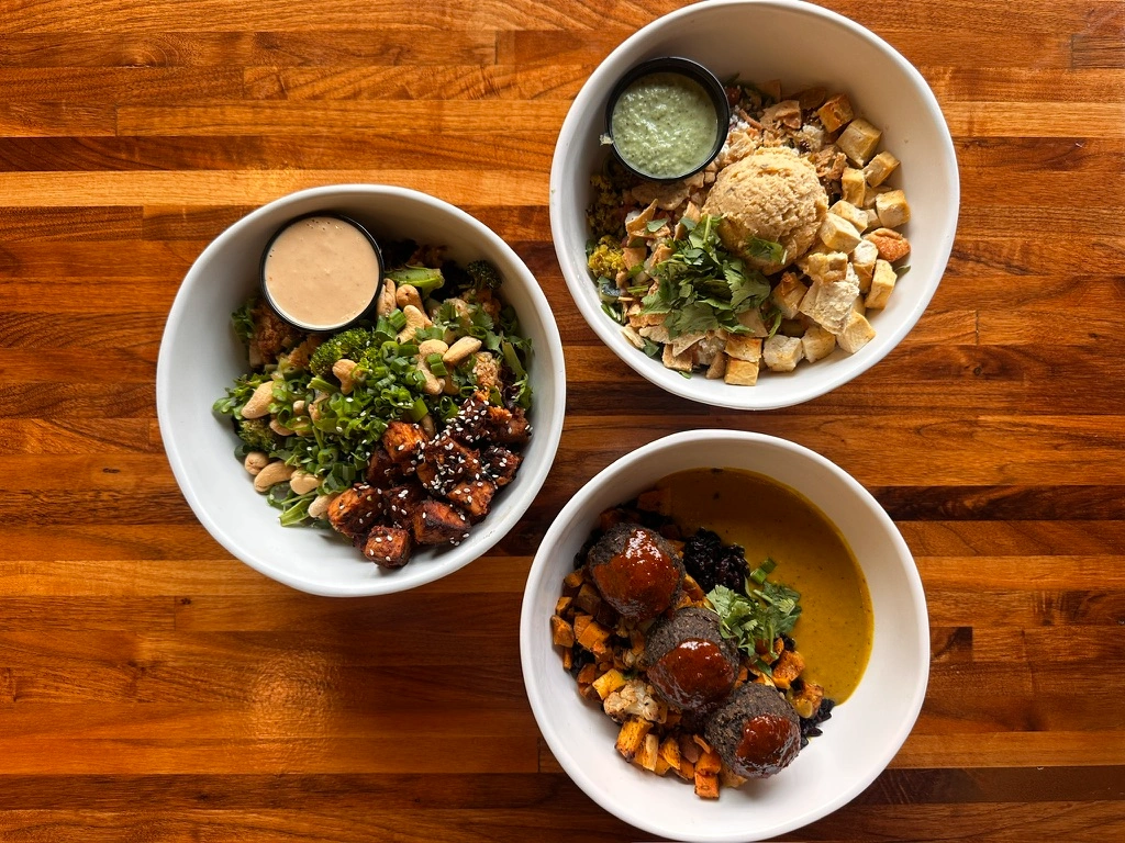 Overhead view of three fresh vegetarian grain and salad bowls with dressings at Forage Kitchen.