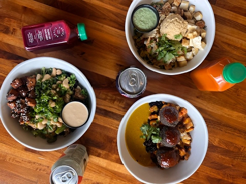 Overhead view of three plant-based grain bowls, juices, and drinks on a wooden table at Forage Kitchen.