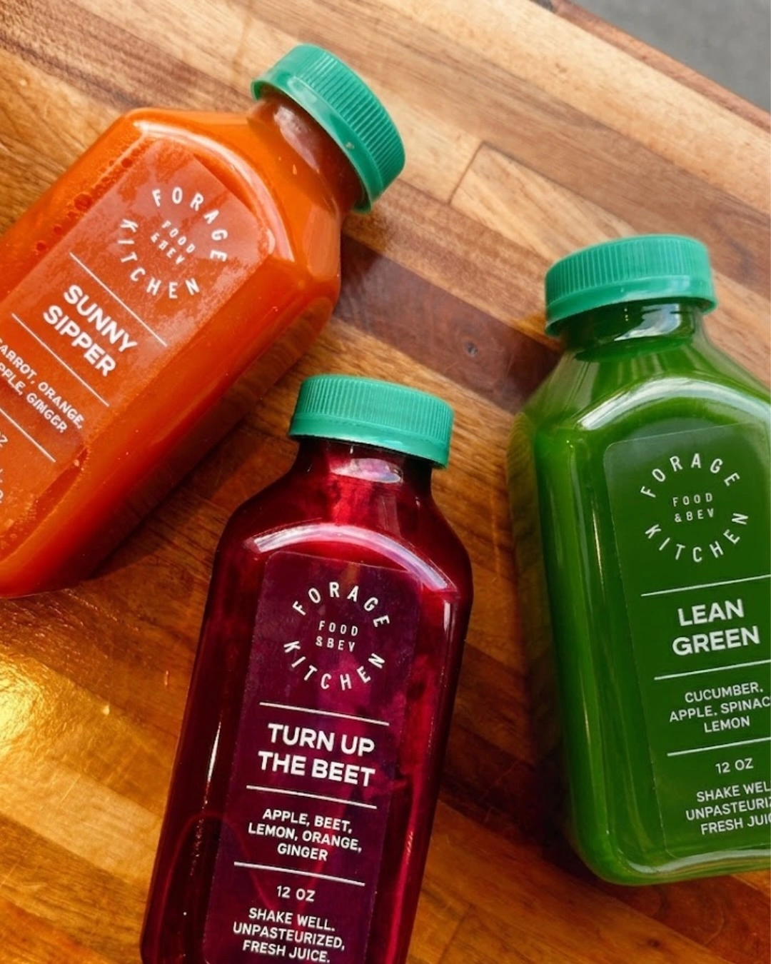 Three different fresh juice bottles: Sunny Sipper, Turn Up The Beet, and Lean Green on a wooden table at Forage Kitchen.