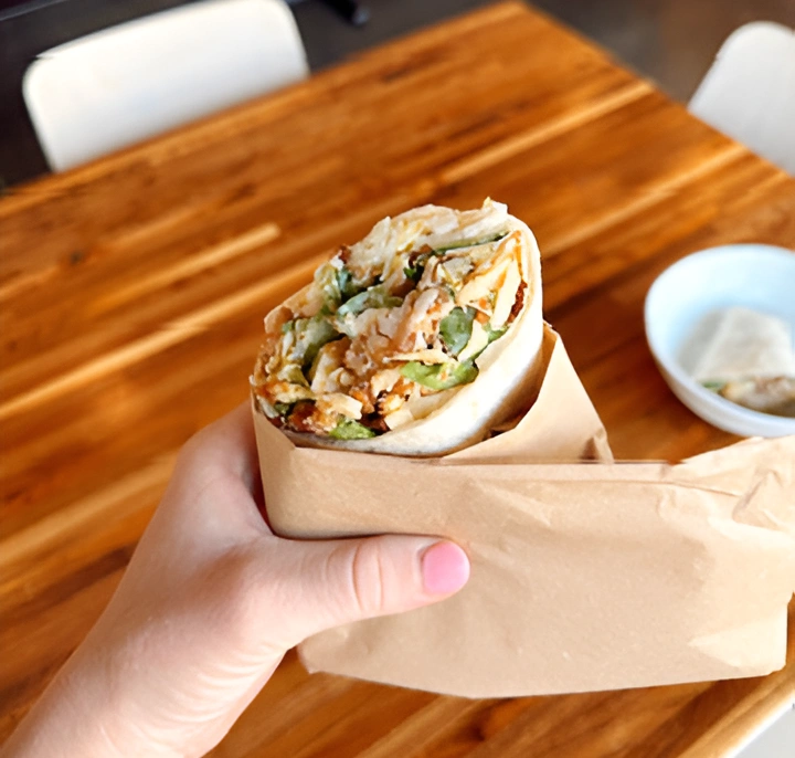 Hand holding a sliced wrap wrapped in brown paper over a wooden table at Forage Kitchen.