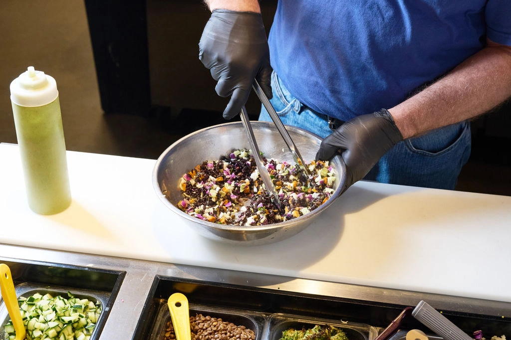 A Forage Kitchen staff member using tongs to mix grain bowl ingredients in a metal bowl.