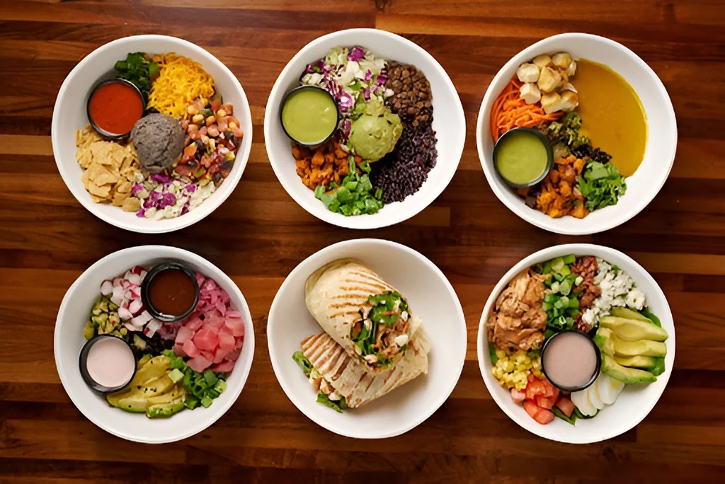 An overhead layout of six different grain bowls, salads, and wraps from Forage Kitchen on a wooden table.