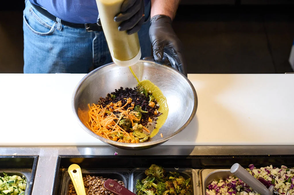 A staff member in black gloves squeezing Thai Basil dressing into a metal bowl at Forage Kitchen.