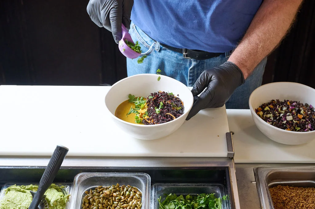 A staff member sprinkling fresh herbs onto Forage Kitchen’s Thai grain bowl with a purple measuring scoop.