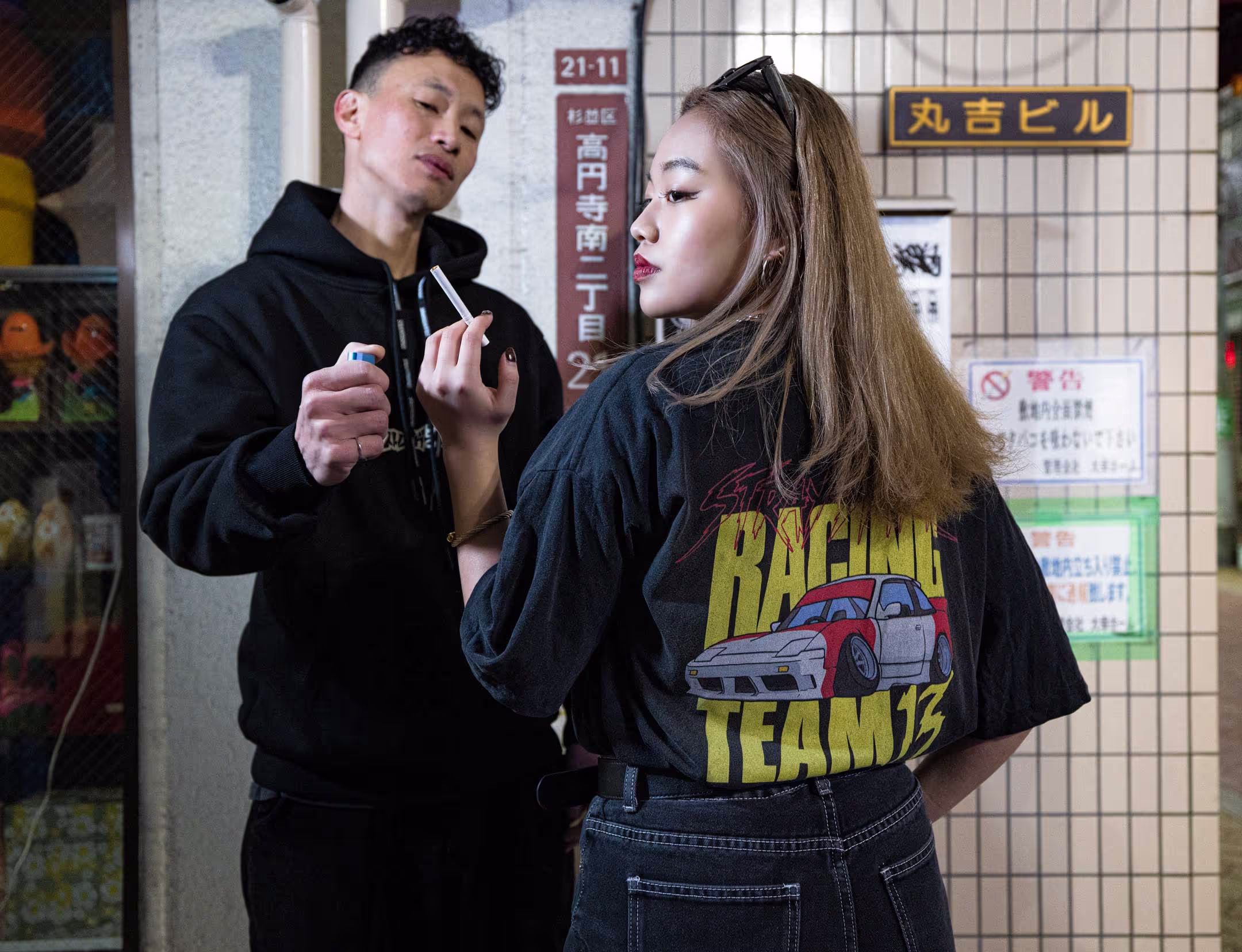 Two people in the streets of Tokyo wearing Straygang branded clothing. The most visible piece of clothing is a black wash, oversized t-shirt, showcasing the vibrant and colorful back print.