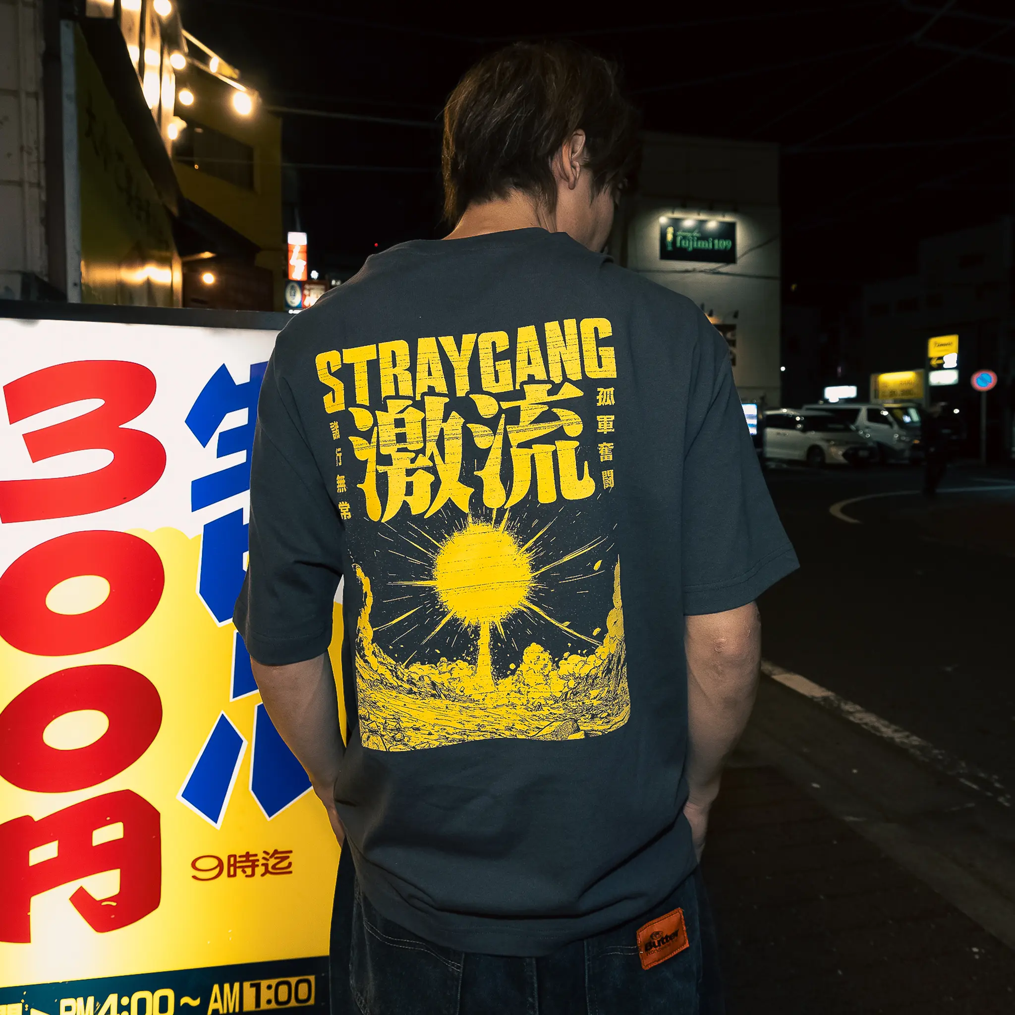 Image that shows off the back design of the grey and yellow supernova tee, worn amidst the back-alleys of Tokyo by a local skateboarder. The large, bright screen print shows an exploding star matched with some loud looking kanji characters.