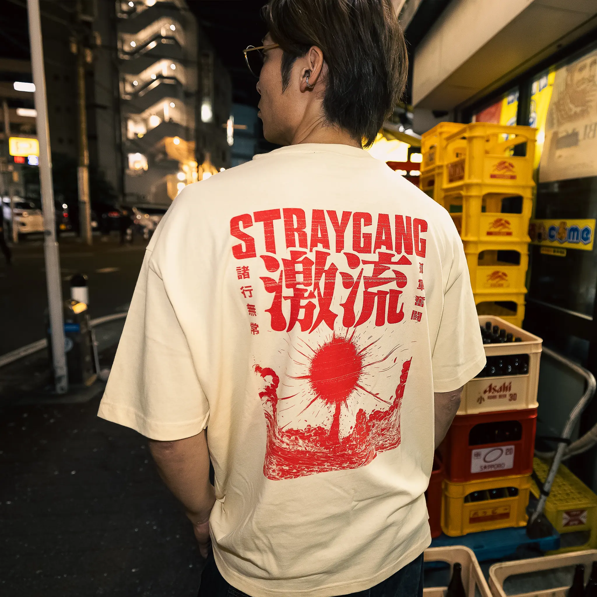Image that shows off the back design of the cream and red supernova tee, worn amidst the back-alleys of Tokyo by a local skateboarder. The large, bright screen print shows an exploding star matched with some loud looking kanji characters.