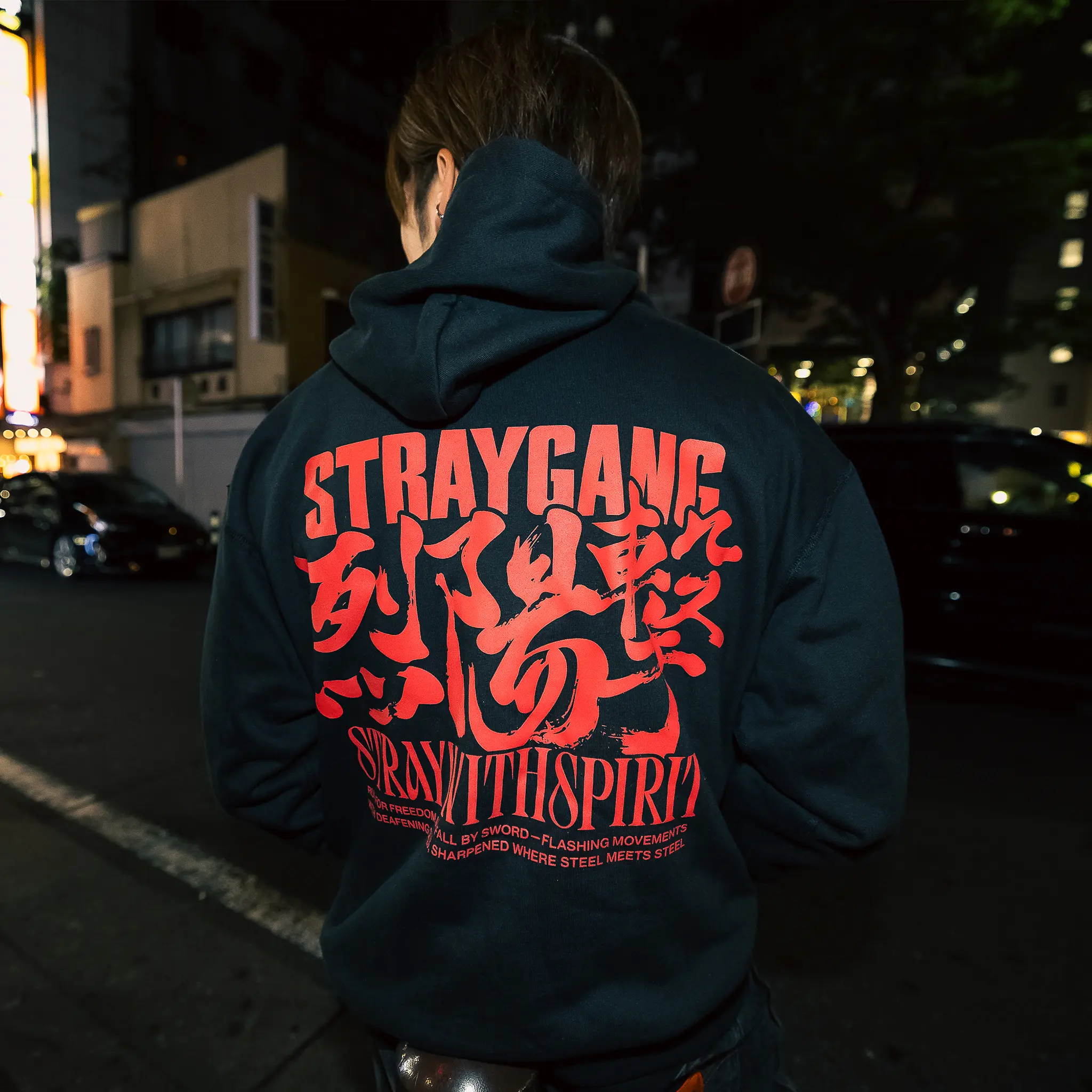 Campaign image of a local skater wearing Straygang's black strike hoodie in the gritty streets of Tokyo. The back design, a large, aggressive screen print matching english with Japanese kanji, mixing two cultures tastefully.