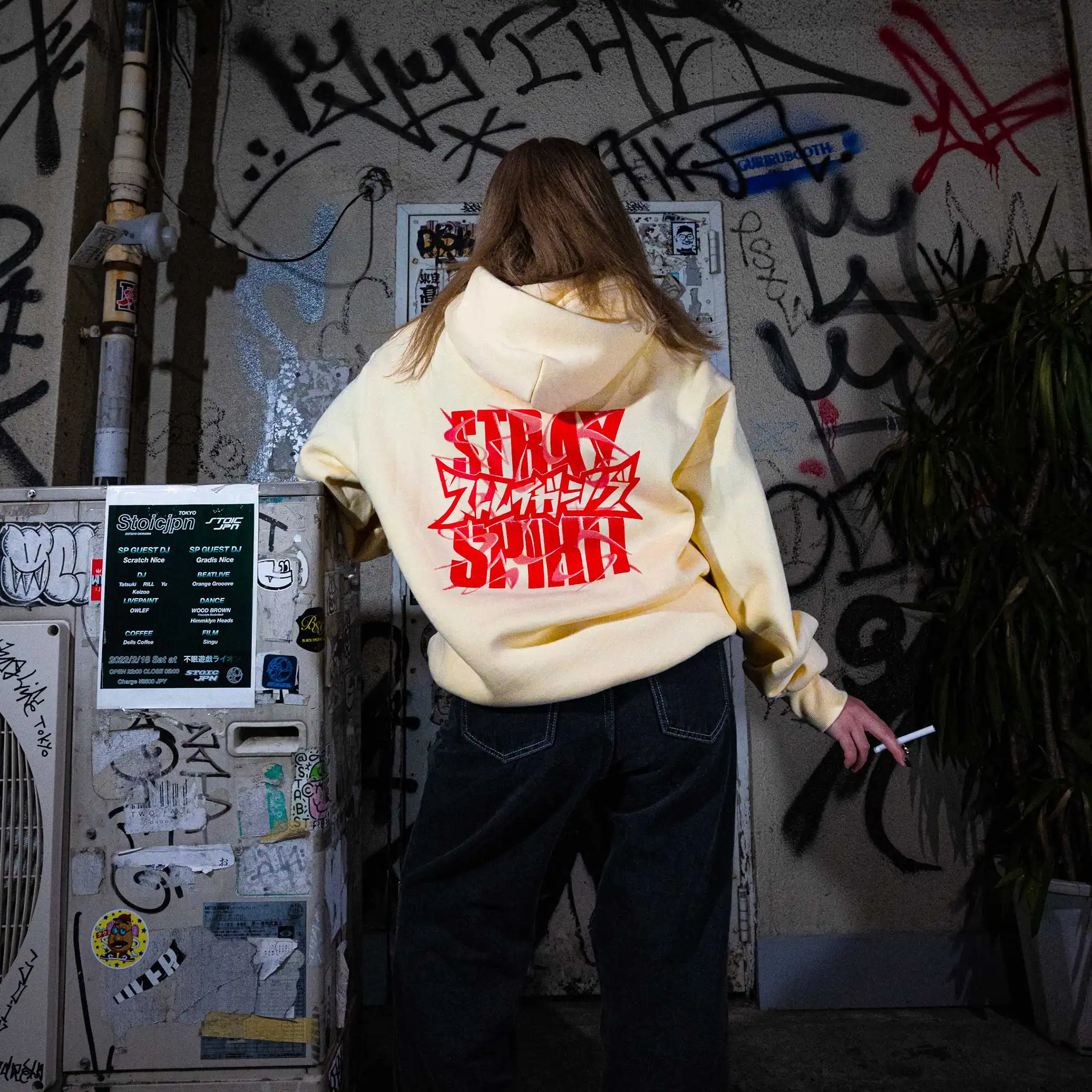 Model in Tokyo back alley wearing Straygang Spirit Hoodie in cream, back view, with large red graphic print.