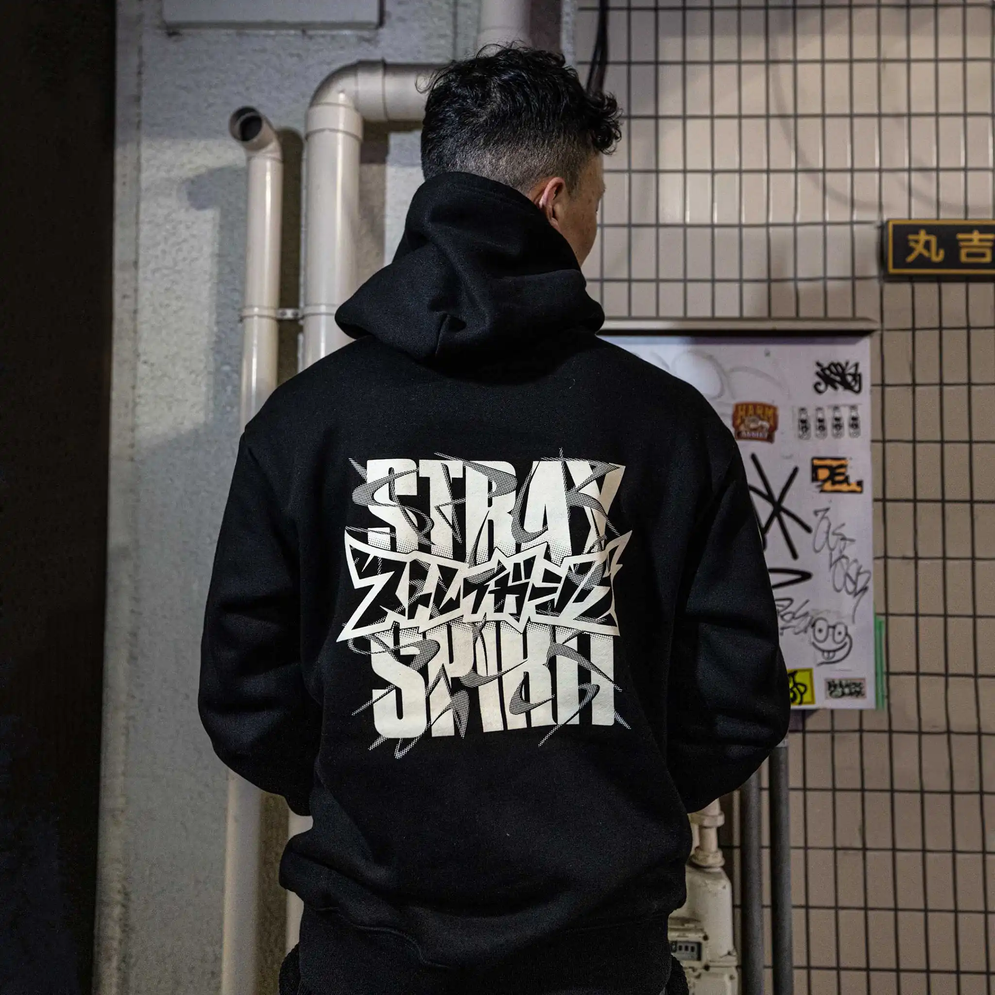 Model wearing Straygang Spirit Hoodie in black, back view, featuring oversized white graffiti-style Katakana print, photographed in Tokyo alley setting.