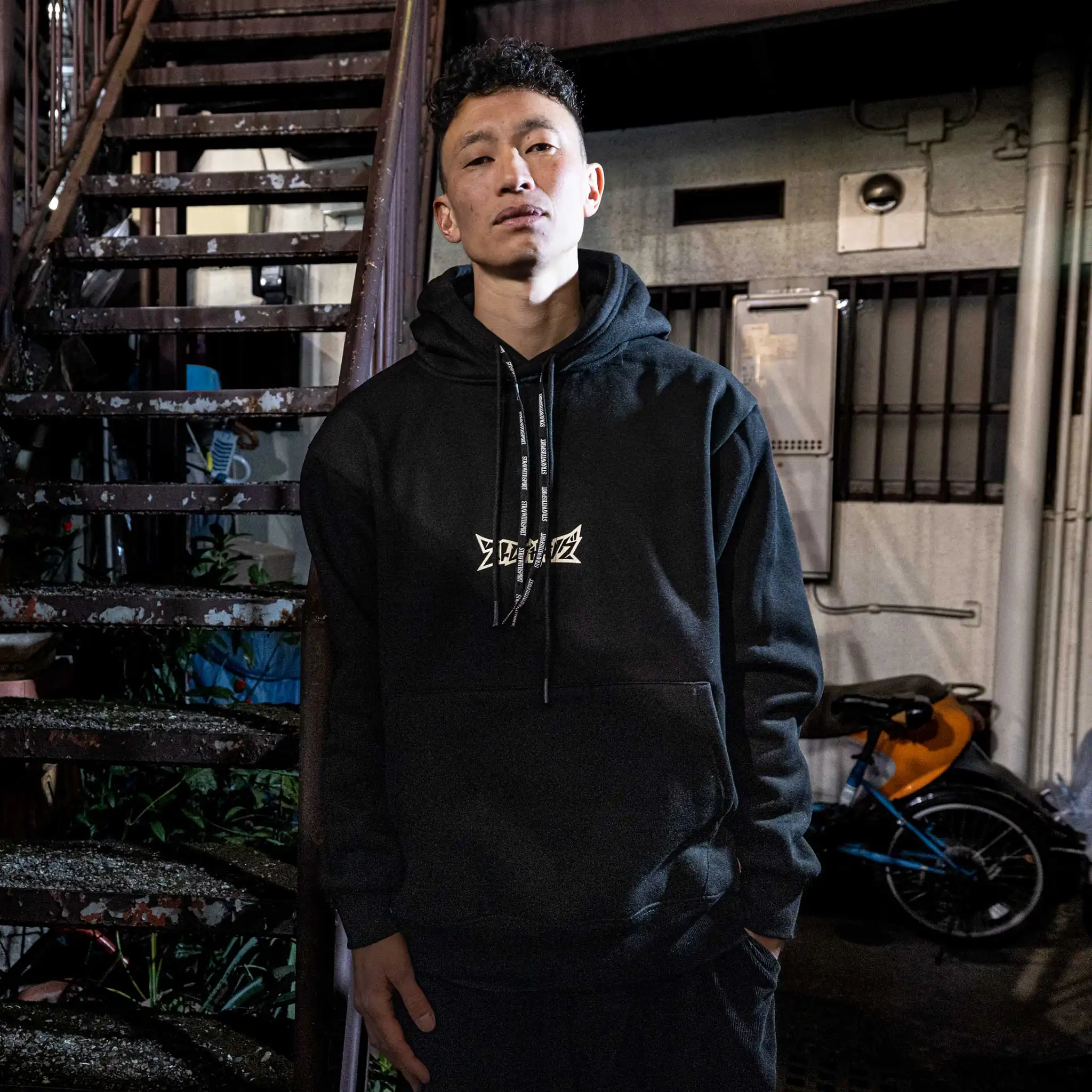 Model wearing Straygang Spirit Hoodie in black, front view, with bold chest logo, shot in Tokyo alleyway at night.