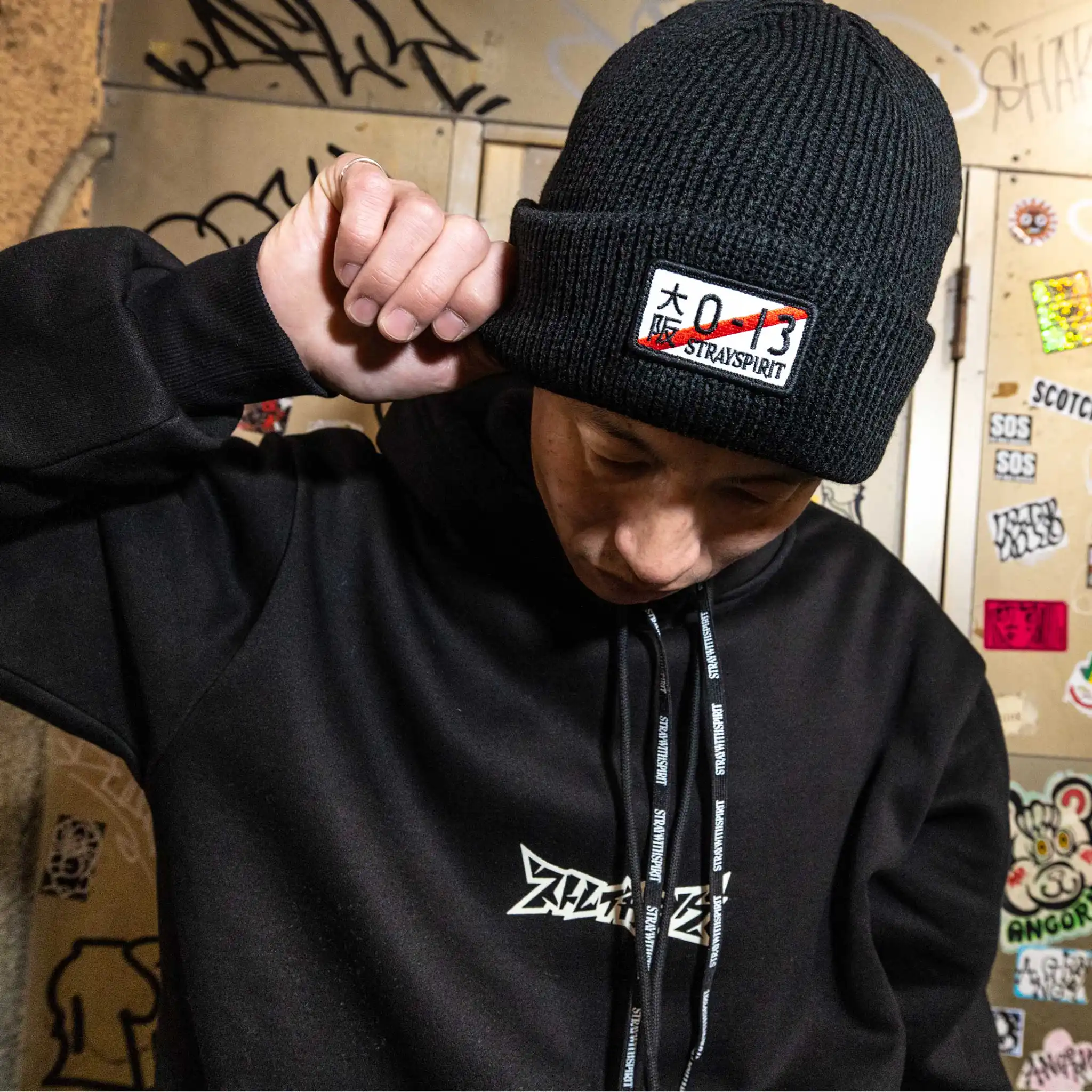 Campaign image highlighting the front design of Straygang's 'Illegal' black beanie, modeled in a Tokyo alley. The custom embroidered patch creates a visual contrast against the backdrop of the dirty Tokyo alley.