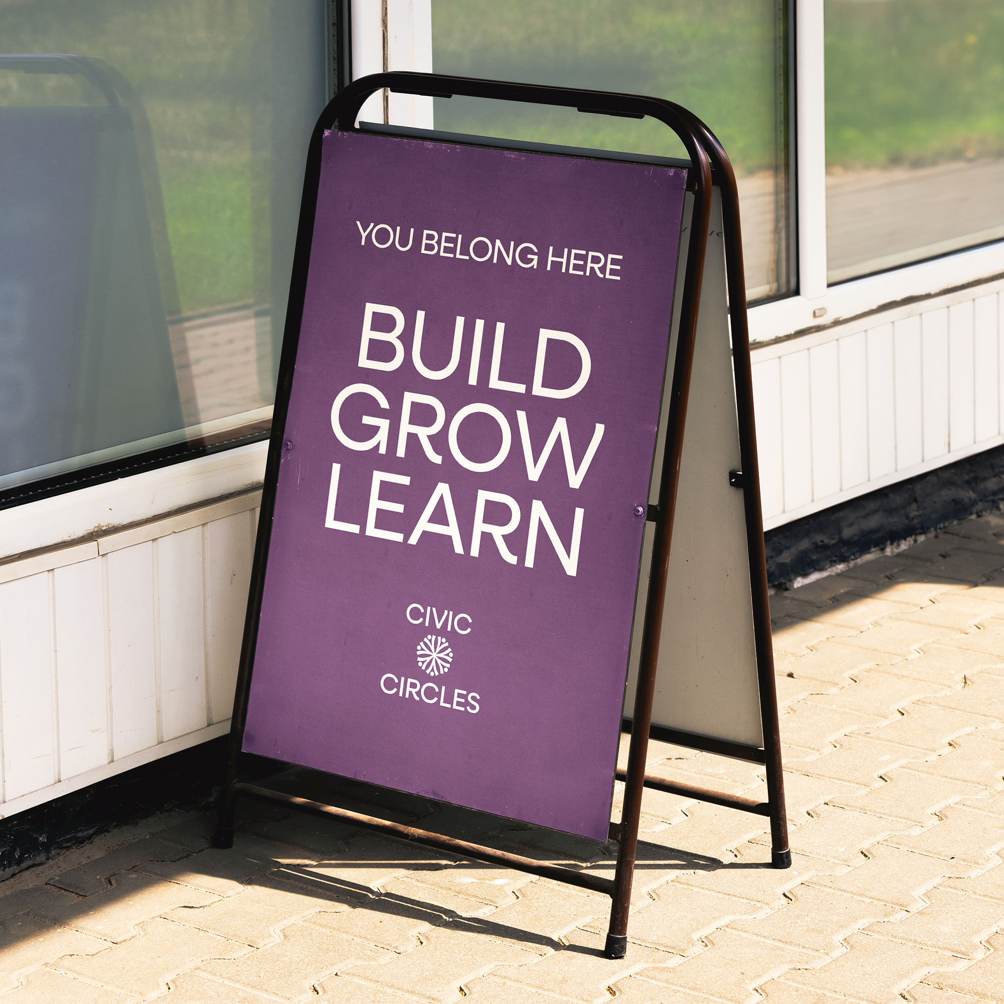 A design mockup showing an A-frame sign in front of a city store on a sidewalk that says, "You belong here. Build, grow, learn."
