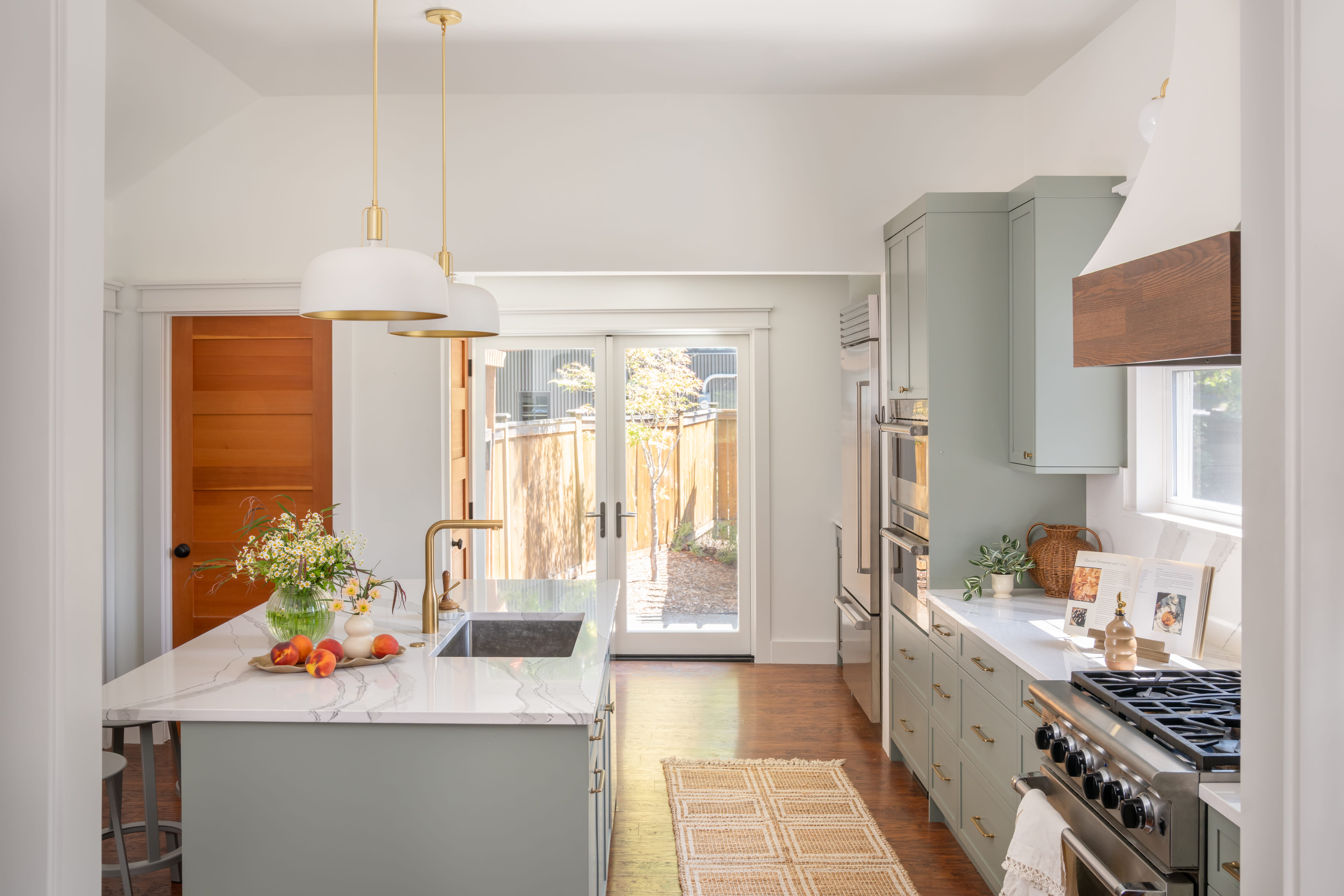 **Alt text:** Modern Bellevue kitchen remodel featuring a large quartz island with an undermount sink, sage green shaker cabinets, brass fixtures, white countertops, a gas range, double wall ovens, and glass patio doors bringing in natural light.
