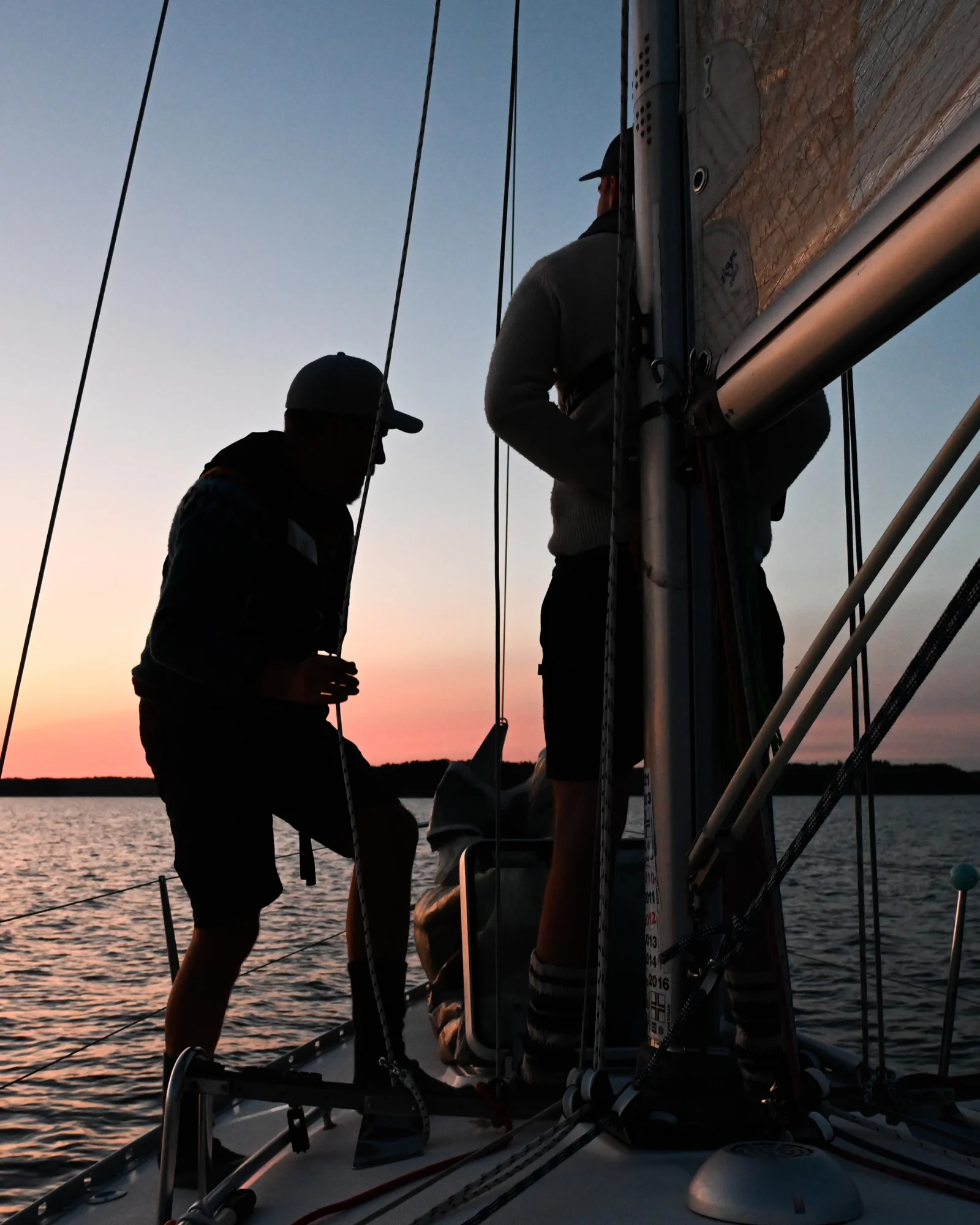 Lähikuva, jossa kaksi henkilöä työskentelee purjeveneen kannella auringonlaskussa.