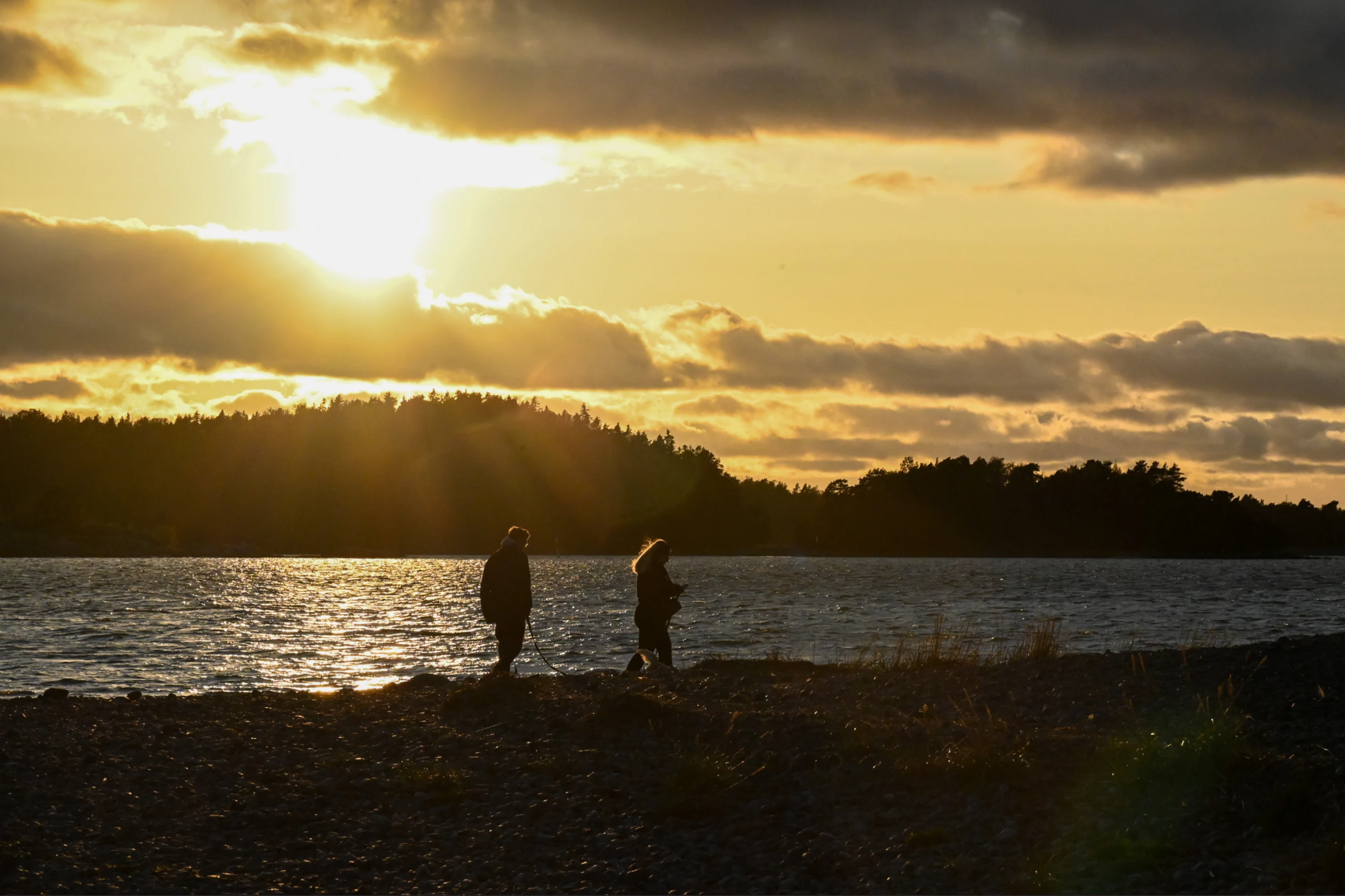 Kaksi henkilöä rannalla auringonlaskussa, taustalla meri ja metsäinen ranta