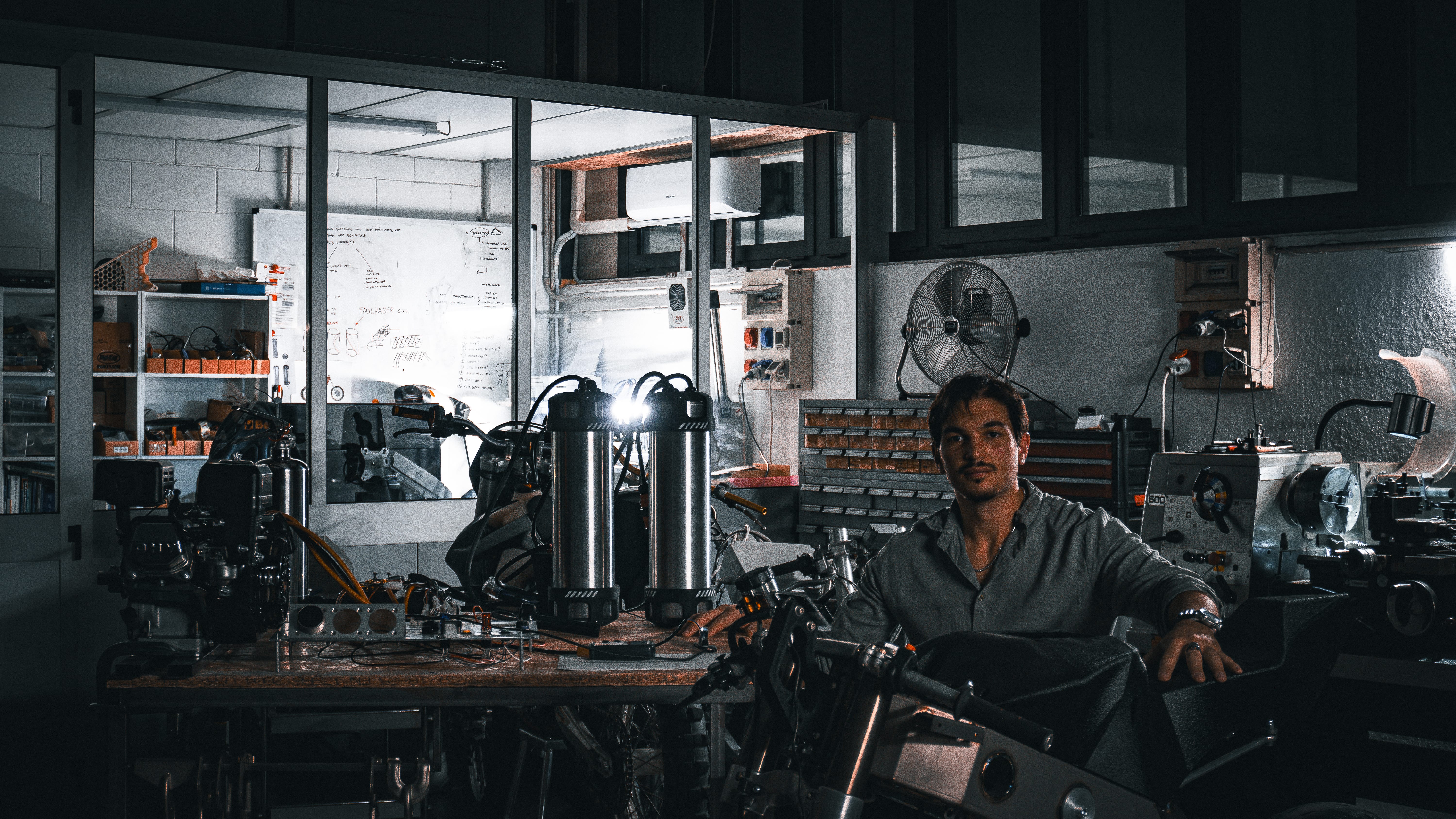 Man sitting in a dimly lit workshop surrounded by mechanical equipment, tools, and motorcycle parts on a wooden workbench with shelves and a fan in the background.