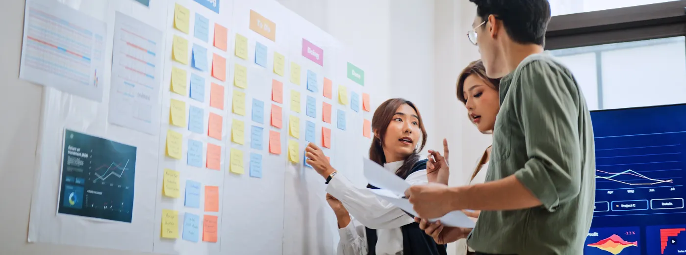 Drei junge Berufstätige diskutieren vor einem Whiteboard mit farbigen Haftnotizen in einem modernen Büro, daneben ein Bildschirm mit Datenvisualisierungen.