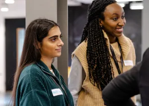 Two women standing indoors during a social or professional event, one wearing a green jacket and the other in a beige vest, both with name tags and engaged in conversation.