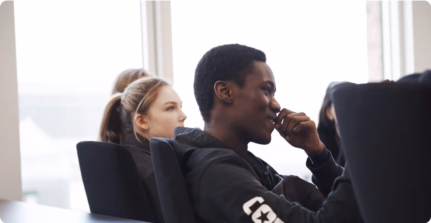 A young man sitting in a conference room with a thoughtful expression, surrounded by other people in the background.