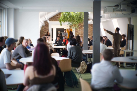 Wide shot of a classroom with students sitting at tables, listening to a presentation