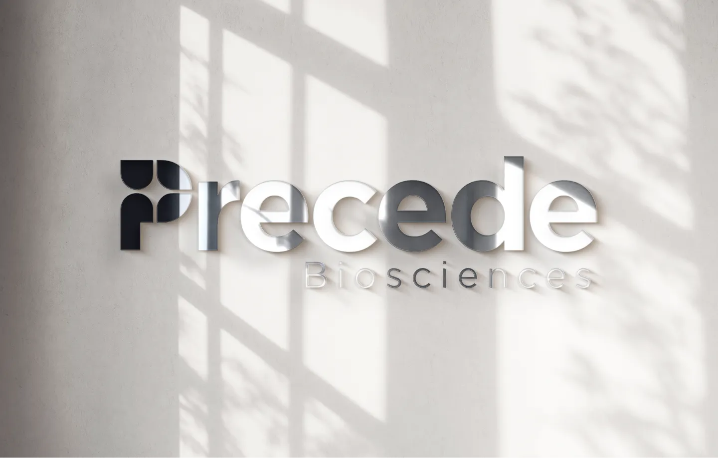 Three-dimensional metallic logo reading 'Precede Biosciences' cast on a wall with shadows from window panes and foliage.
