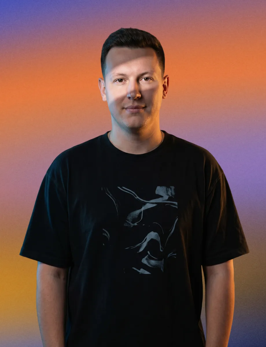 Man in a black t-shirt standing against a gradient background of orange, purple, and yellow hues.
