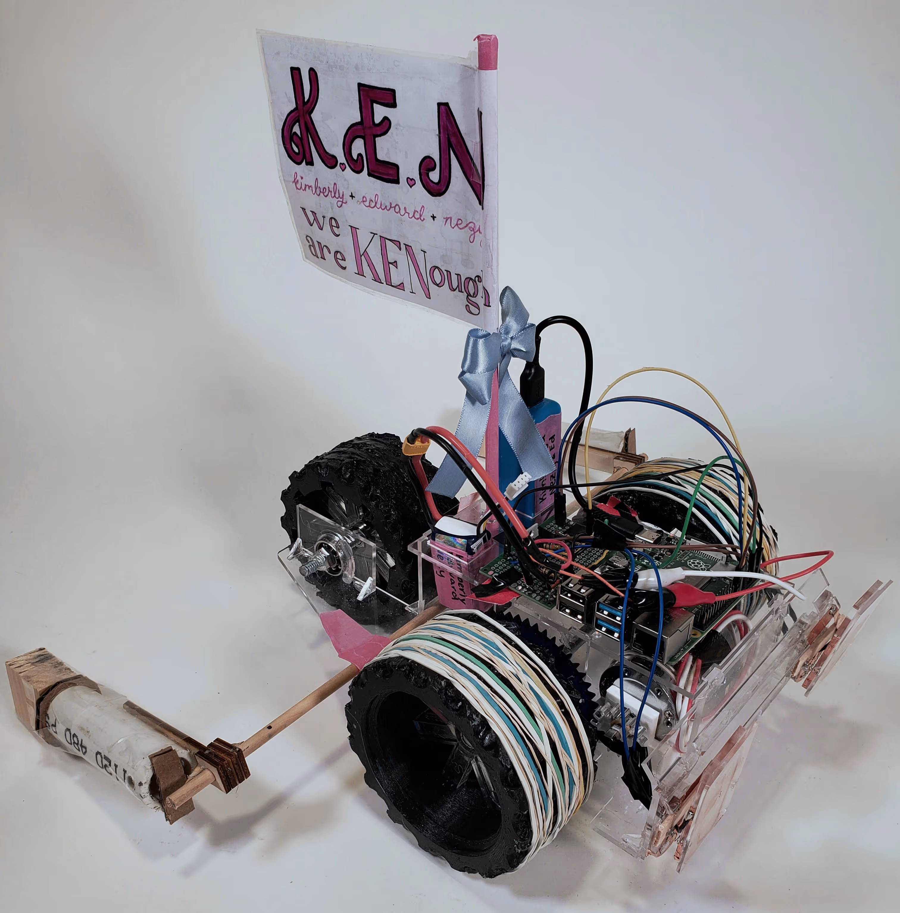 The KEN robot car with three large monster truck wheels with added rubber bands for traction, a raspberri pi and protoboard with wires sticking out on top of the bed and a guiding beam towards the back of the car.