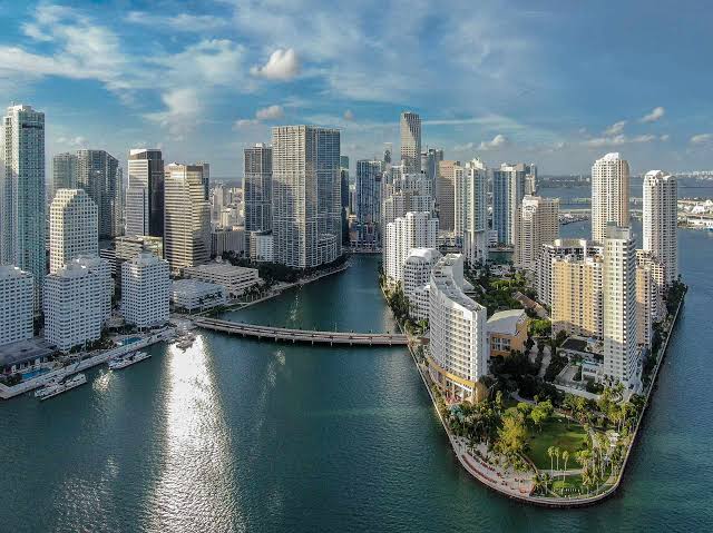 Aerial view of a city waterfront with high-rise buildings along a curved shoreline and a bridge over water under a blue sky with scattered clouds.