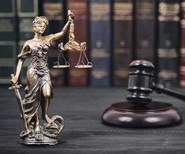 Bronze statue of Lady Justice holding scales and a sword, with a judge's wooden gavel and law books blurred in the background.