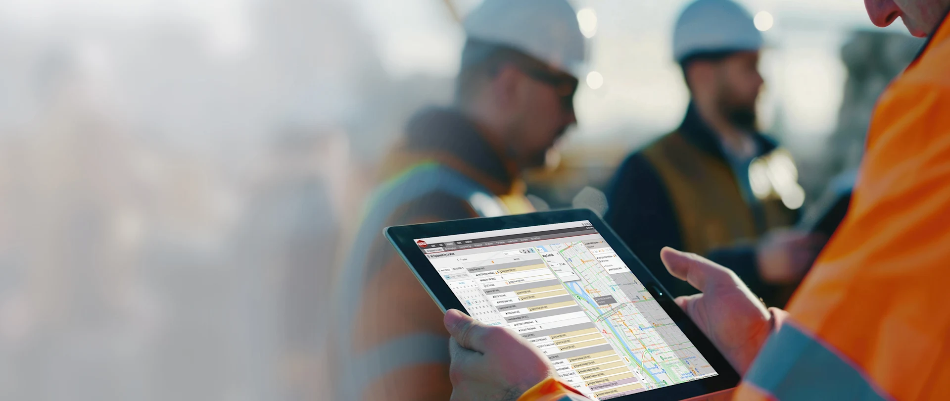 A person in an orange construction jacket holds a tablet displaying a digital schedule and map interface, with two blurred construction workers in white hard hats in the background.